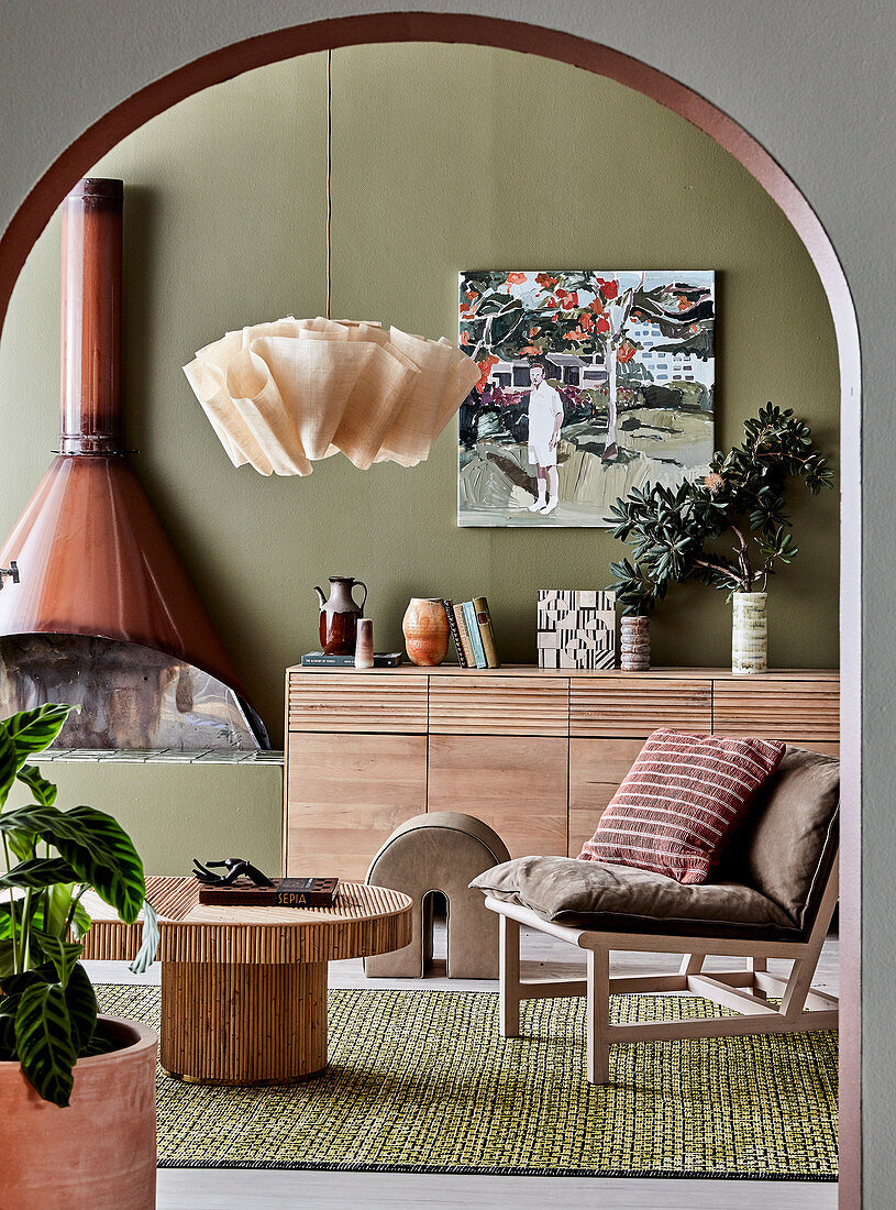 View through round arch on designer rattan table, chair, sideboard and fireplace in the living room with olive green wall