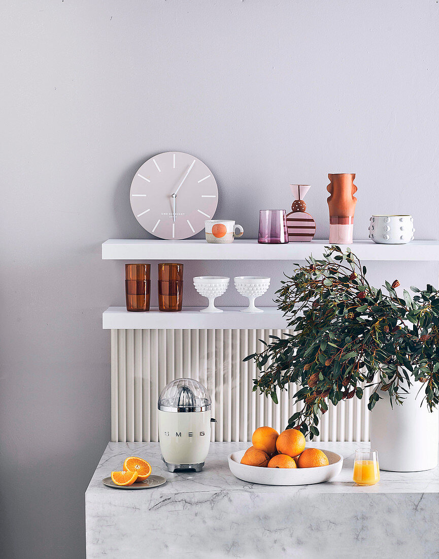 Marmortheke mit Zitruspresse, Orangen und Blätterzweig in Vase, im Hintergrund Regalbretter mit Uhr und Geschirr