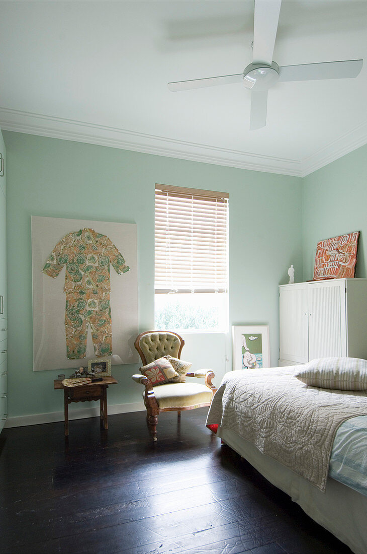 Bett, weißer Schrank, Polsterstuhl vor Fenster und Kinderkleidung als Wanddekkoration in pastellgrünem Zimmer