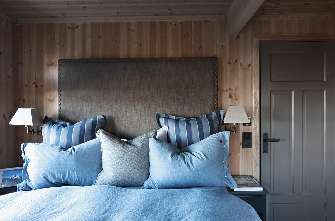 Bed with grey headboard and blue bed linen against wooden wall in cottage bedroom