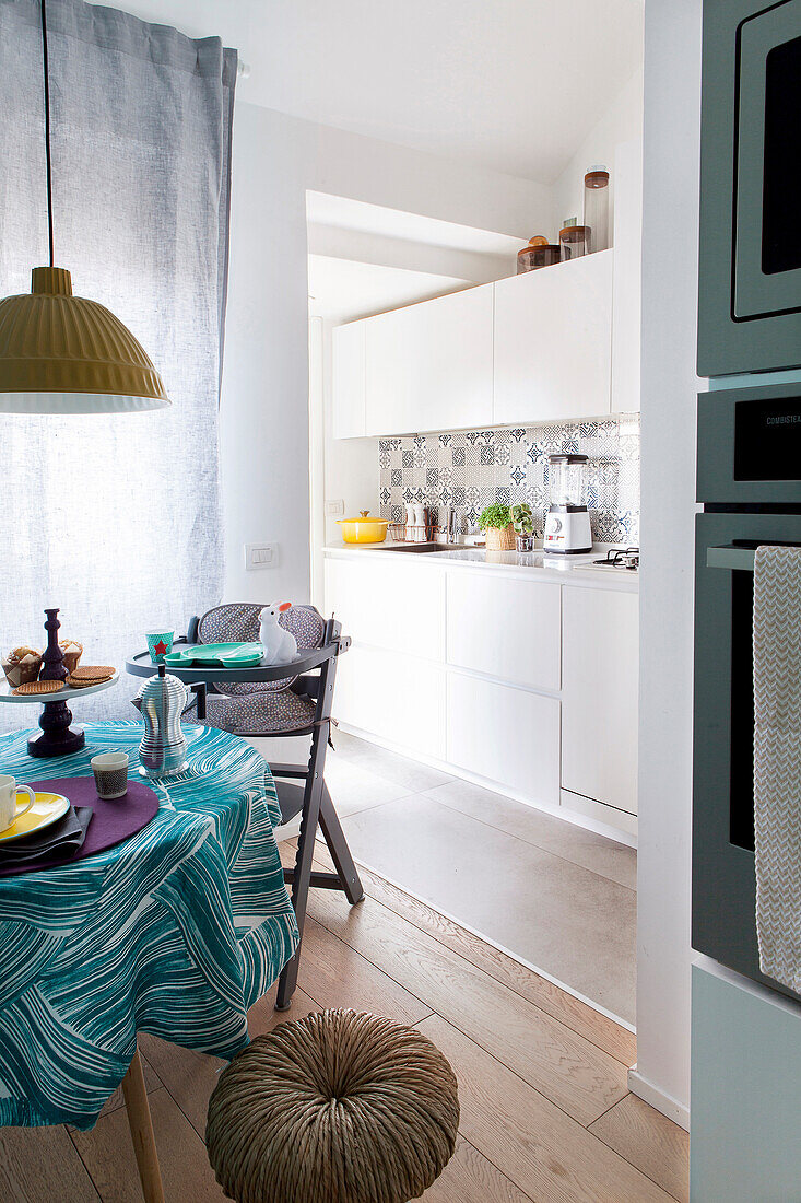 Round table with turquoise tablecloth, stool and highchair and view into white fitted kitchen