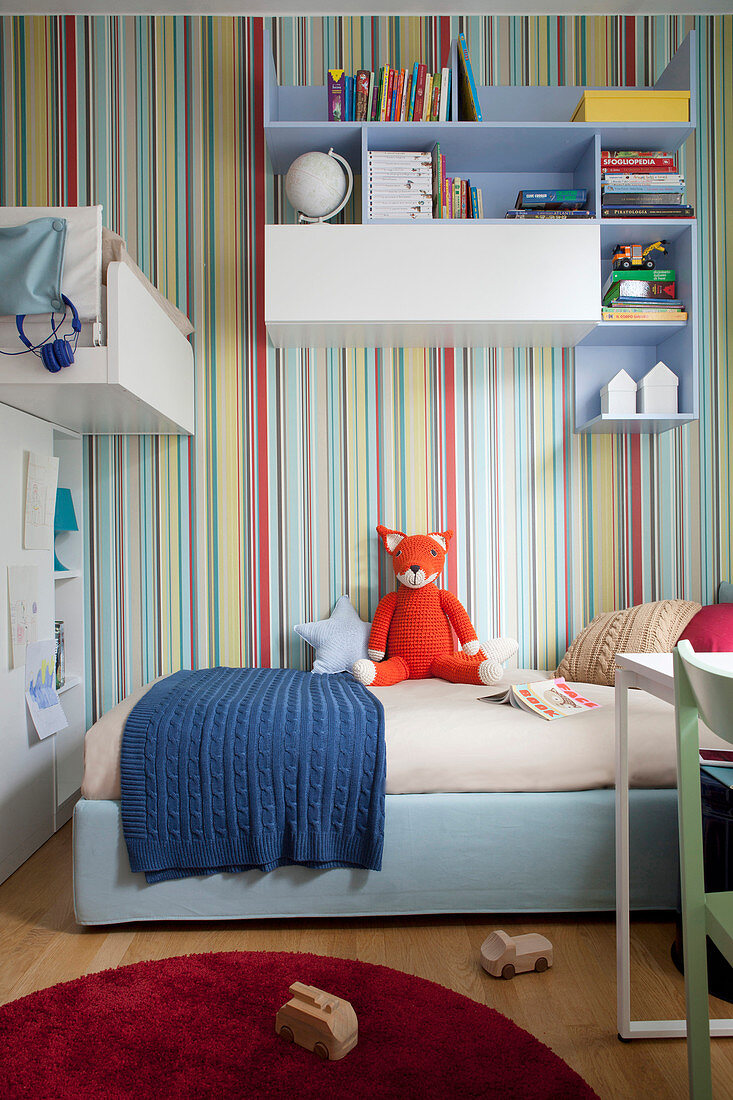 Bed below wall-mounted shelves on multicoloured striped wallpaper in sibling's bedroom