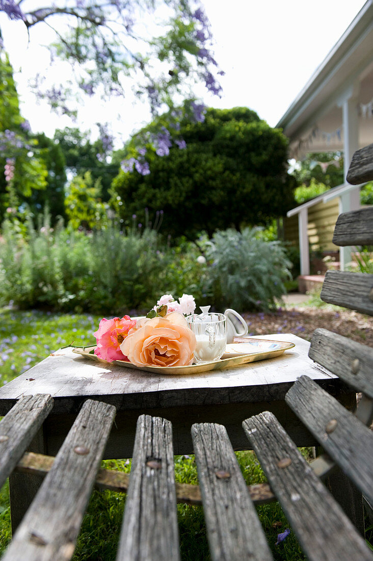 Tablett mit romantischen Rosenblüten auf Holztisch neben verwitterter Holzbank im Garten