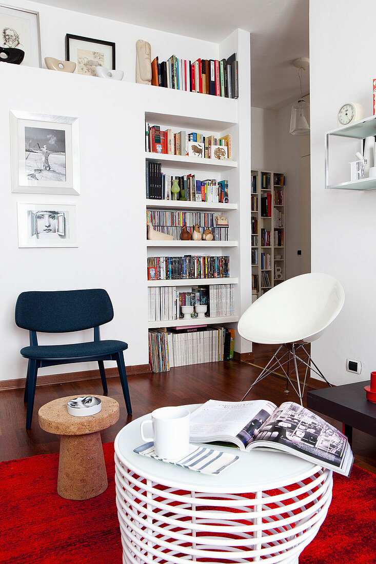 Wohnraum mit weißen Wänden, Einbauregal, verschiedenen Stühlen und rotem Teppich als Farbakzent