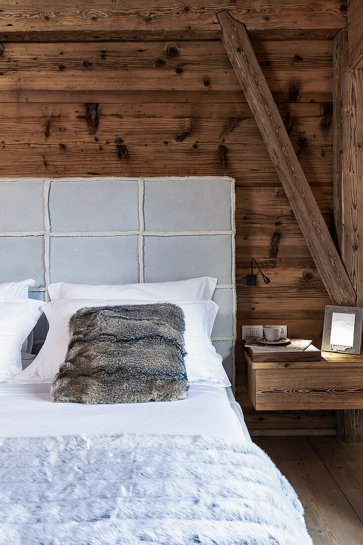 Bett mit gepolstertem Betthaupt und Fellkissen vor rustikaler Holzwand