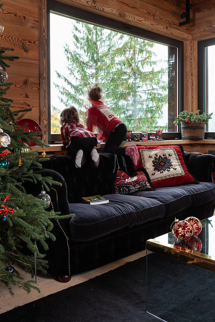 Mädchen auf schwarzem Polstersofa am Fenster in weihnachtlich dekoriertem Wohnzimmer eines Chalets