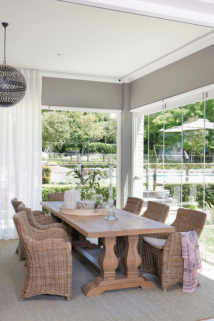 Wuchtiger Holztisch mit Korbstühlen vor Fensterfront mit faltbaren Glastüren und Blick in den Garten