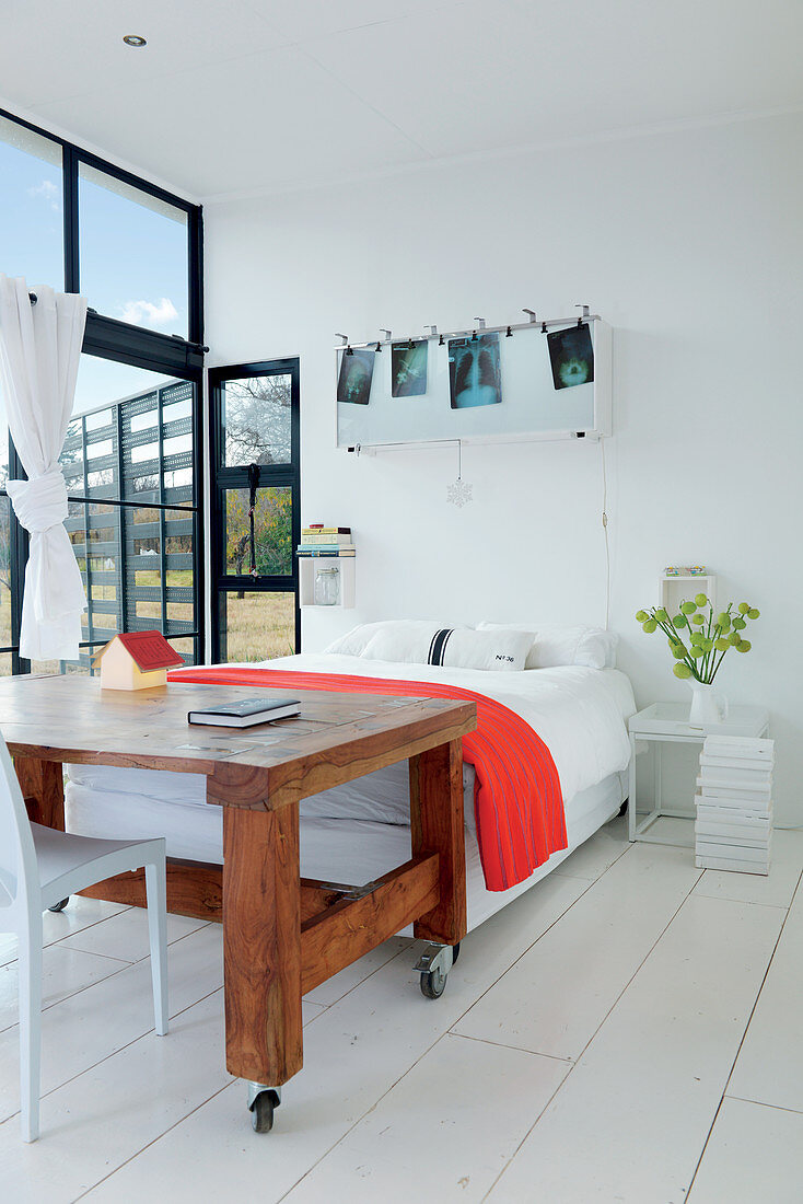 Bett und rollbarer Holztisch in lichtdurchflutetem Wohnraum neben Stahlrahmen-Fensterfront