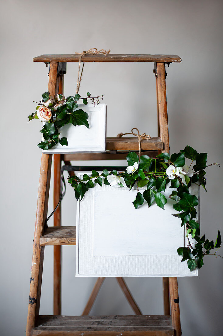 Efeuranken und Stoffblumen um weiß bemalte Bilder an einer Leiter