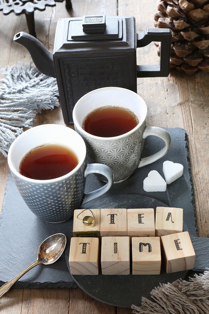 Holzwürfel mit der Aufschrift 'Teatime' auf Schieferplatte mit Teekanne und Teetassen