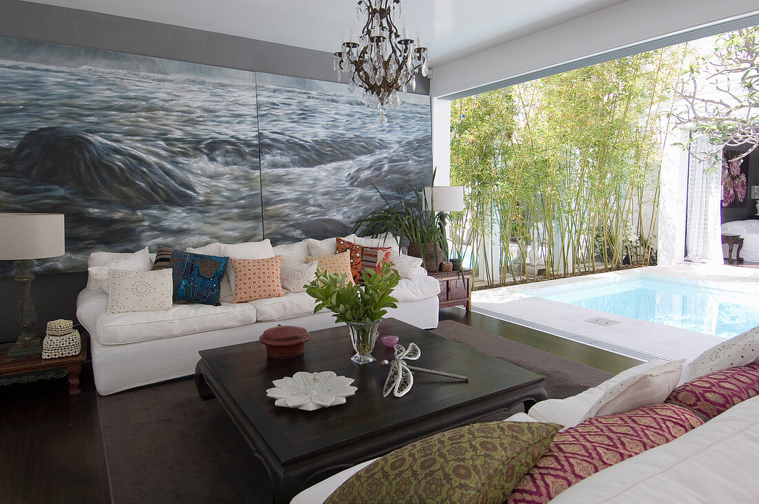 Coffee table and sofas with scatter cushions in front of maritime mural wallpaper; view of pool on terrace