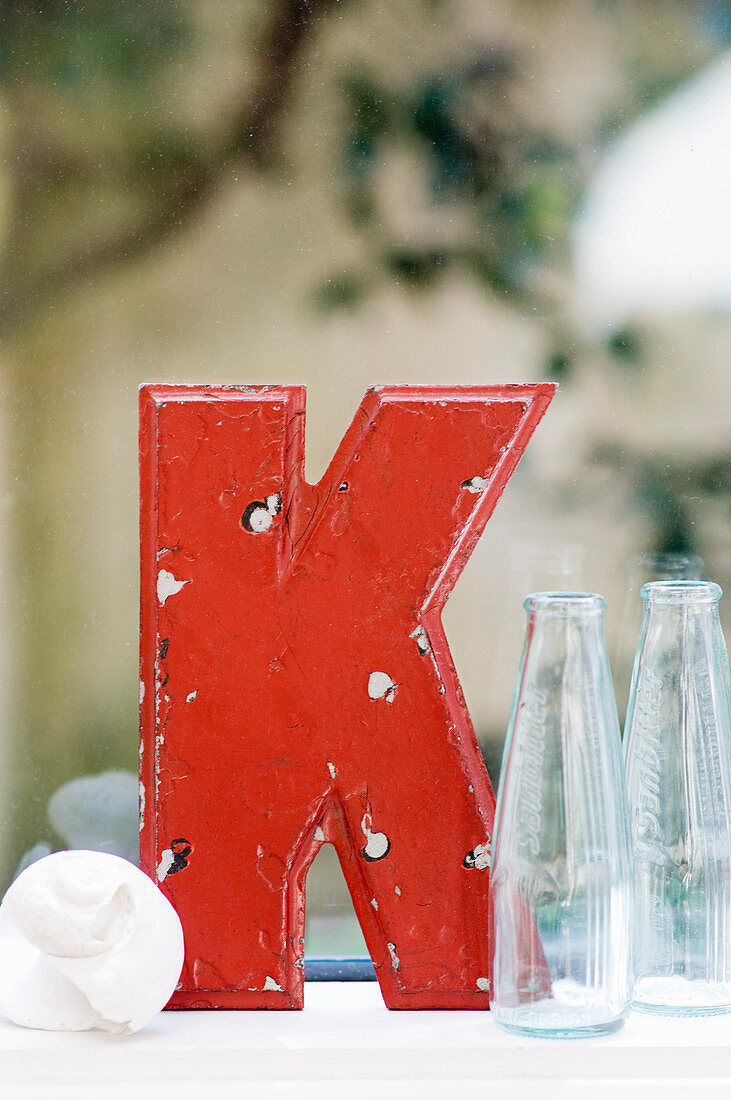 Roter Vintage-Dekobuchstabe K neben Muschel und Glasflaschen