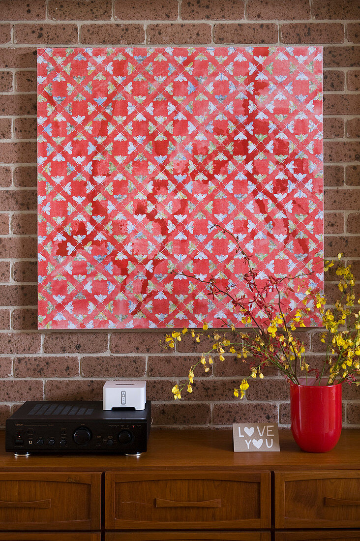 Red geometric artwork on brick wall above wooden chest of drawers