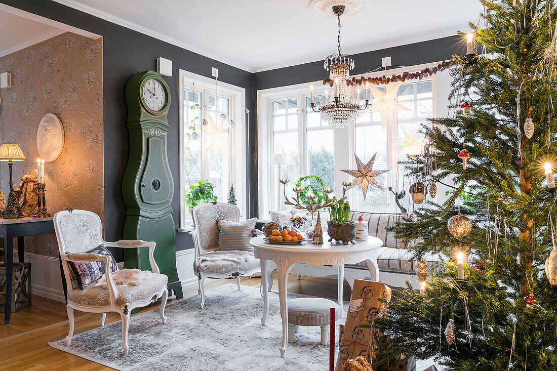 Runder Tisch, darüber Kristallleuchter, Sitzmöbel, Standuhr und beleuchteter Weihnachtsbaum im Salon