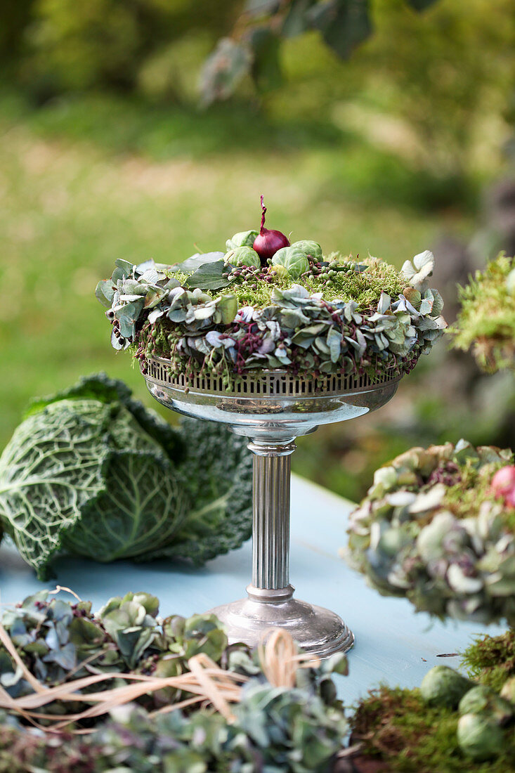 Autumn wreath made of moss, hydrangea blossoms, Brussels sprouts, and onions on a silver bowl with a base