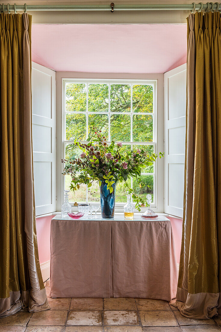 Tisch mit Blumenstrauß vor Fenster mit geöffneten Fensterläden, Vorhänge aus goldener Seid