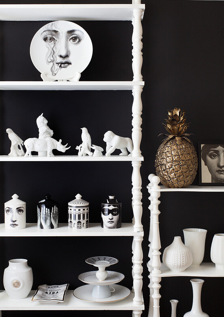 Porcelain figurines and Fornasetti ornaments on shelves against black wall