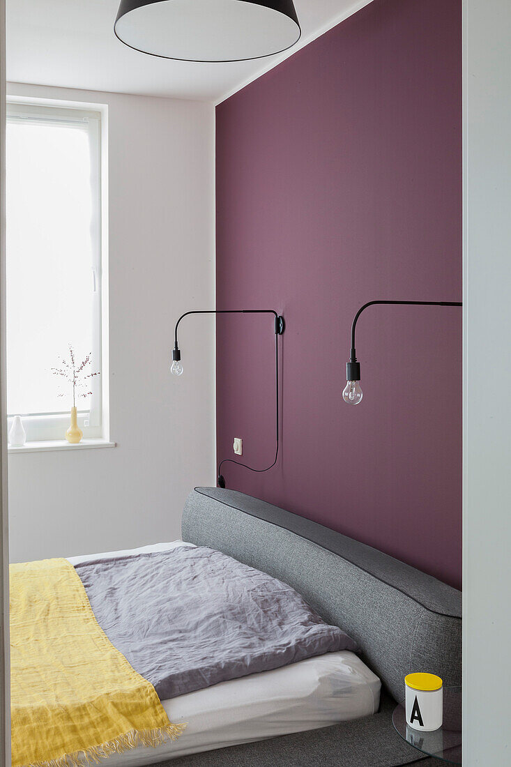 Double bed with grey headboard against plum-coloured wall with reading lamps