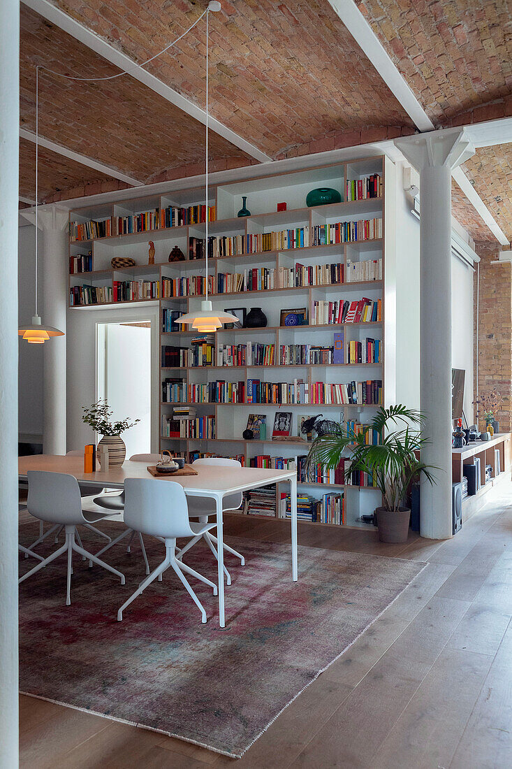 Eßtisch mit Stühlen, darüber Pendelleuchte und Bücherwand in offenem Wohnraum mit hoher Decke