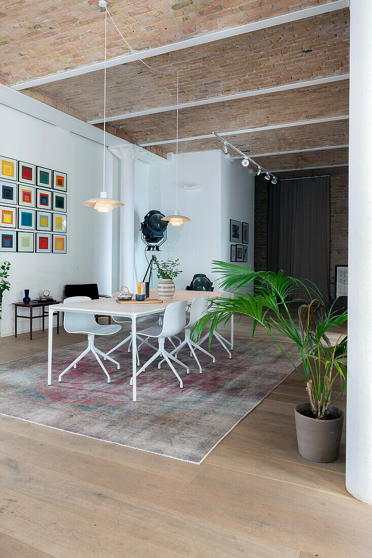 Weißer Esstisch mit Stühlen, darüber Pendelleuchte und bunte Bilder an der Wand in offenem Wohnraum mit hoher Decke