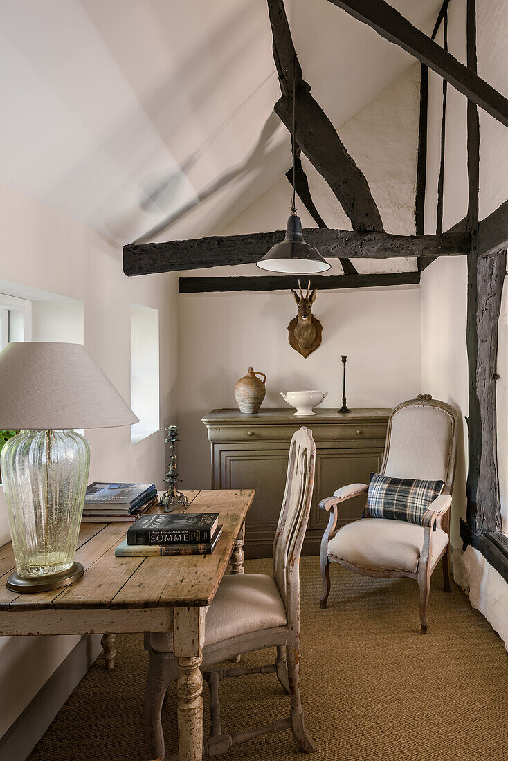 Wooden table with table lamp and upholstered chair in attic room with exposed wooden beams
