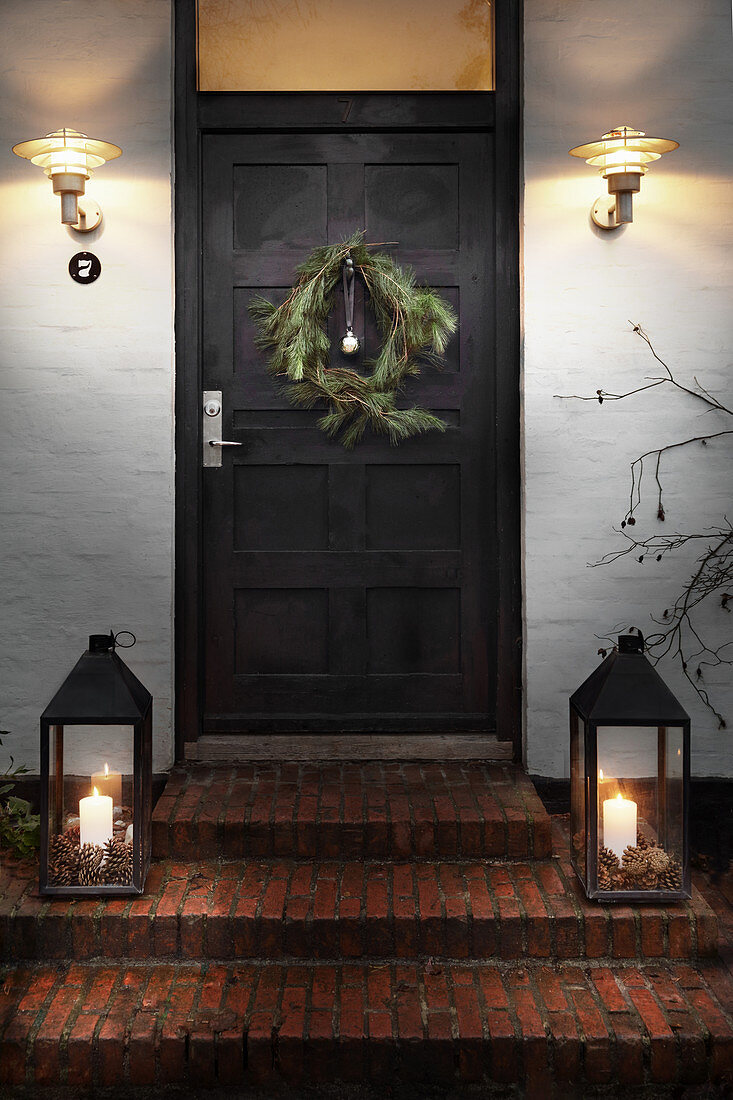 Christmas wreath on front door flanked by wall-mounted lamps and candle lanterns on steps
