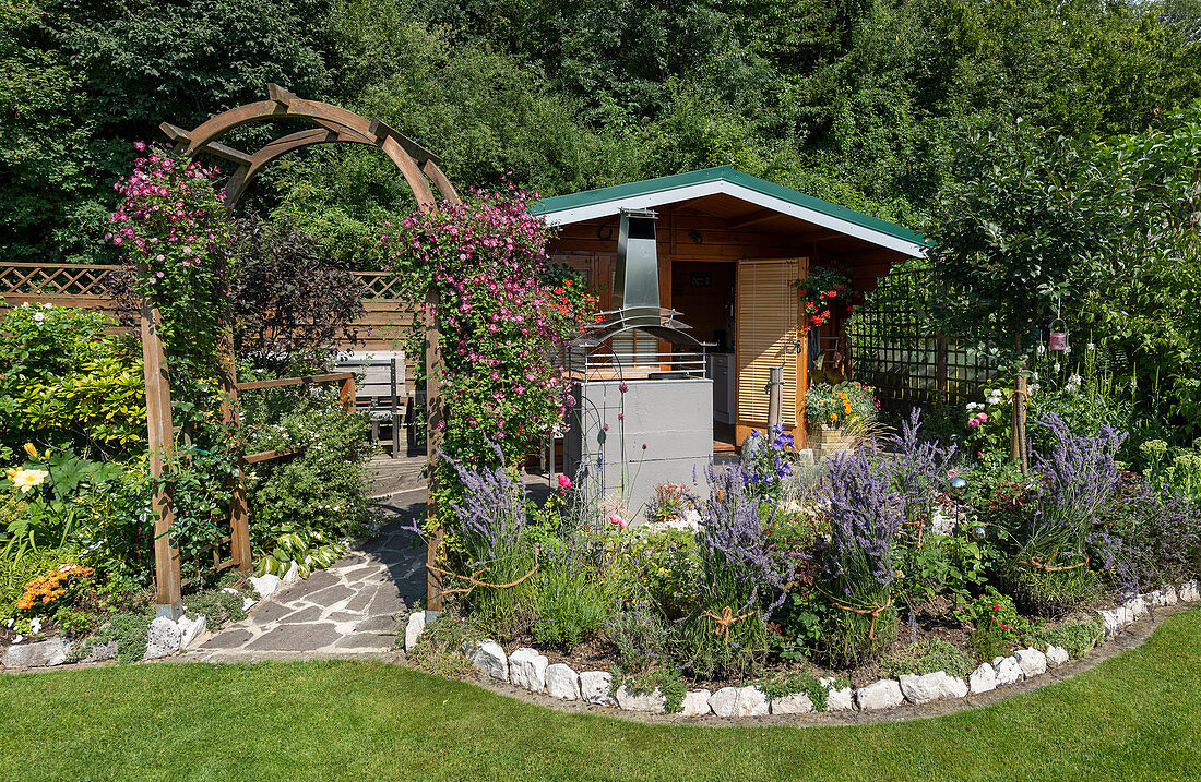 Sommerlicher Schrebergarten mit Gartenhaus, Rosenbogen mit Waldrebe 'Avant-Garde' und Staudenbeet mit zusammengebundenem Lavendel
