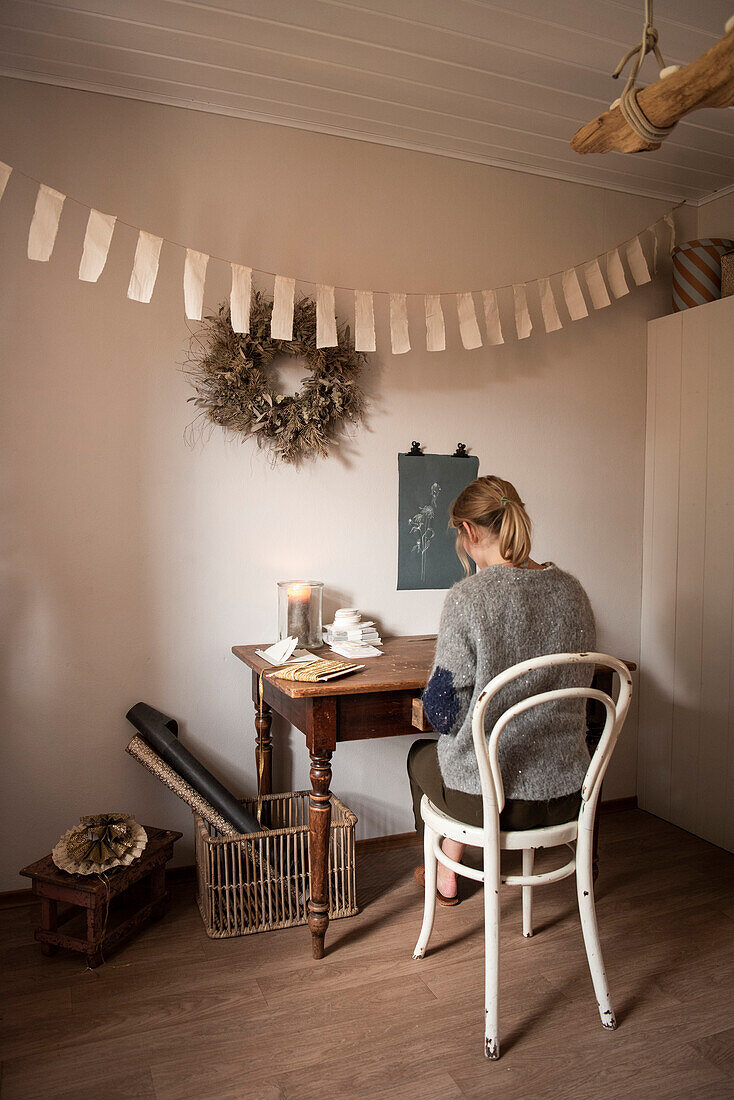 Woman sitting at old wooden table below DIY bunting and wreath on the wall