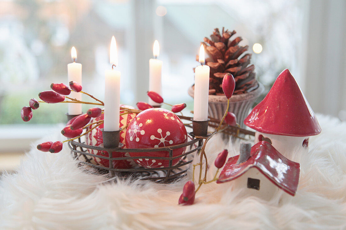 Eisenkorb als Adventskranz mit rot-weißen Christbaumkugeln, Hagebutten, Keramikhäuschen und Zapfen