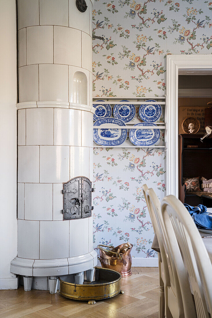Weißer Kachelofen im Zimmer mit gemusterter Tapete und blau-weißen Wandtellern