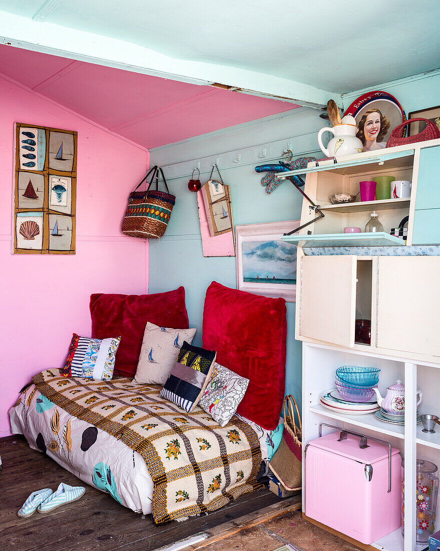 Cushions on a daybed next to a crockery cupboard in a beach hut with pink and blue walls