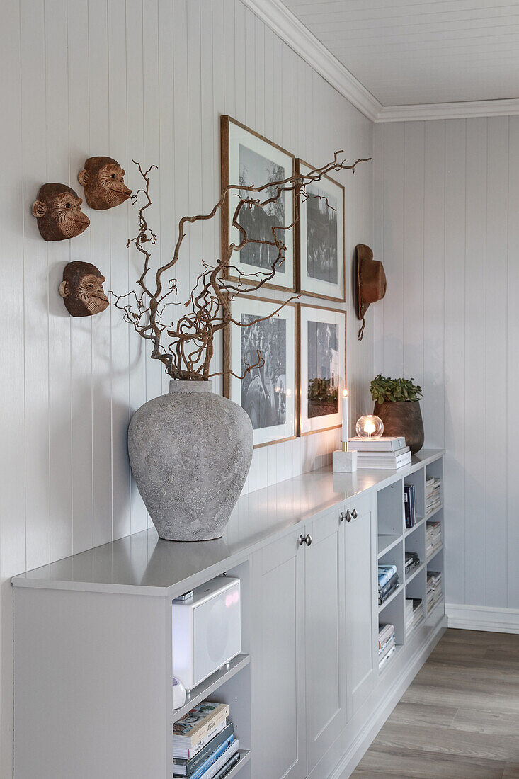 Vase mit Ästen auf Sideboard, darüber Affenköpfe und Fotos im Zimmer