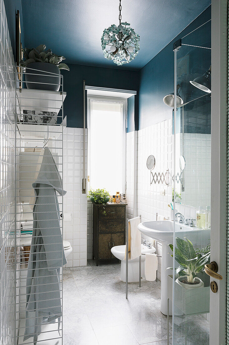 Kleines Badezimmer mit weißen Wandfliesen und blauer Wand, im Vordergrund Regal und Duschbereich