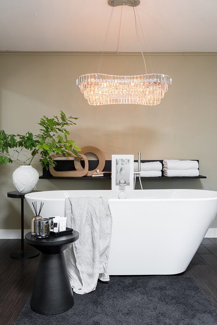 Free-standing bathtub below elegant light fixture in bathroom with beige-coloured wall