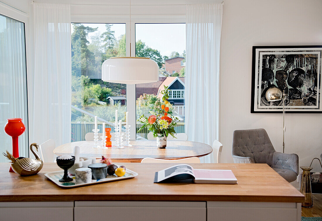 Tray and book on kitchen counter in front of oval dining table with candles next to window
