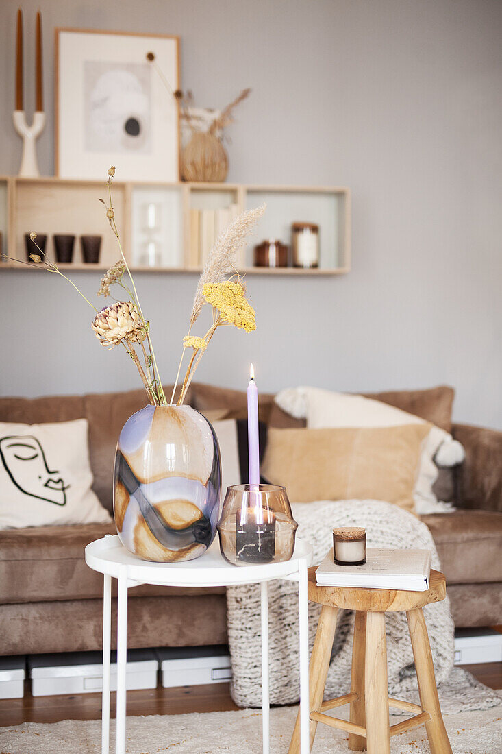 Glass vase of dried flowers, candle and lantern on side table in front of sofa with scatter cushions