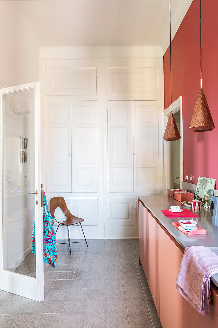 Kitchen base units below pendant lights in front of red wall and and white, floor-to-ceiling, fitted cupboards in the background