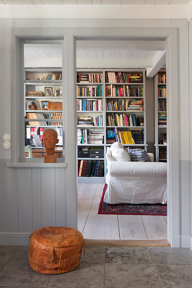 Blick ins Zimmer mit raumhohem Bücherregal und Hussensessel, im Vordergrund Lederpouf