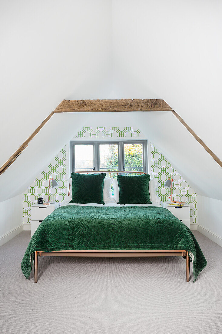 Double bed with green bedspread in attic bedroom with white walls and patterned wallpaper on accent wall
