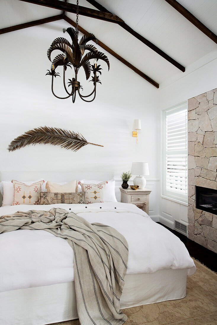 Doppelbett in hellem Schlafzimmer, darüber getrockneter Palmenzweig und Kronleuchter mit Palmenmotiv