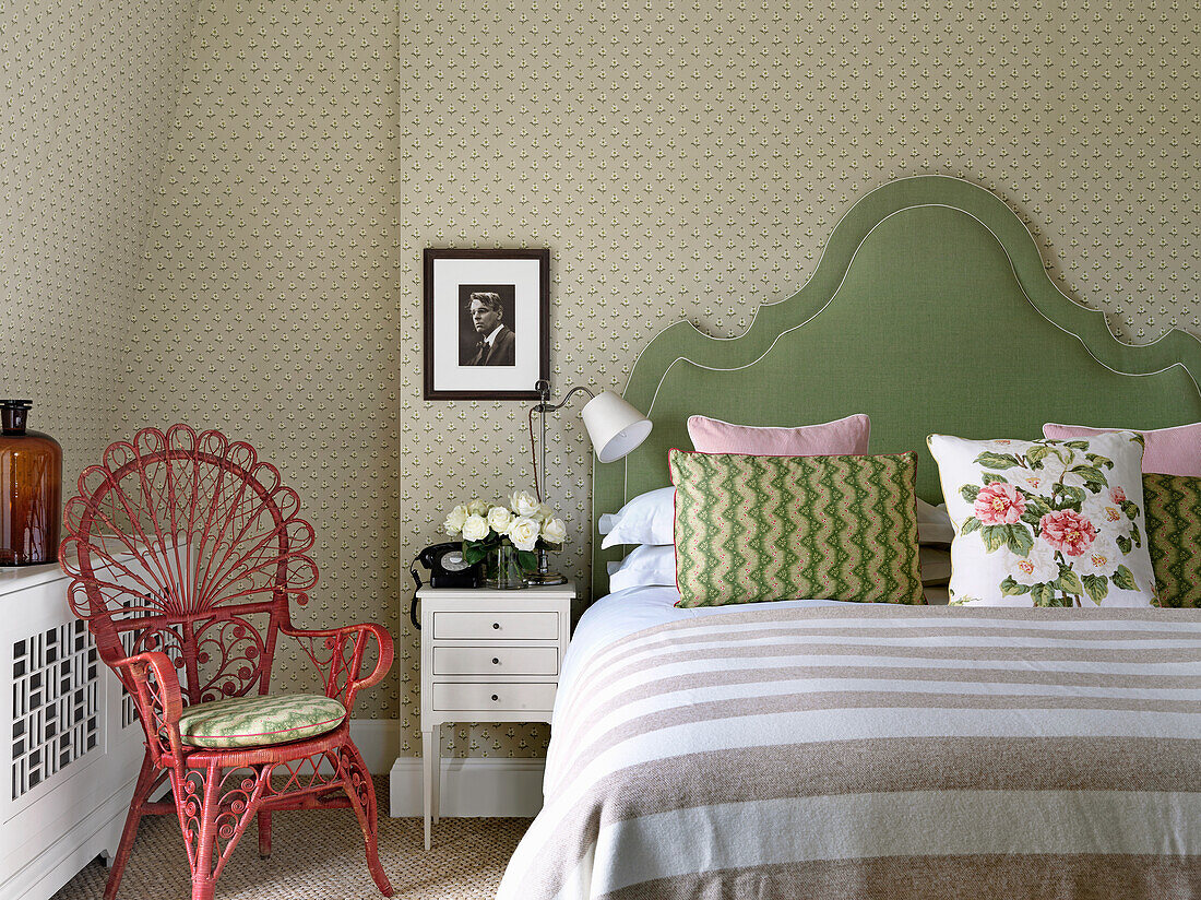 Double bed with green headboard and pretty rattan armchair in bedroom with patterned wallpaper