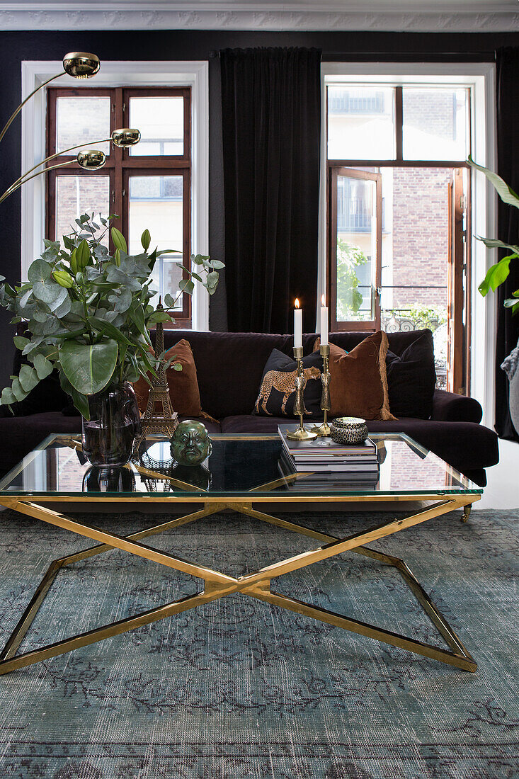 Wohnzimmer mit dunklem Sofa, Couchtisch aus Glas mit goldenem Gestell und dunklen Vorhängen