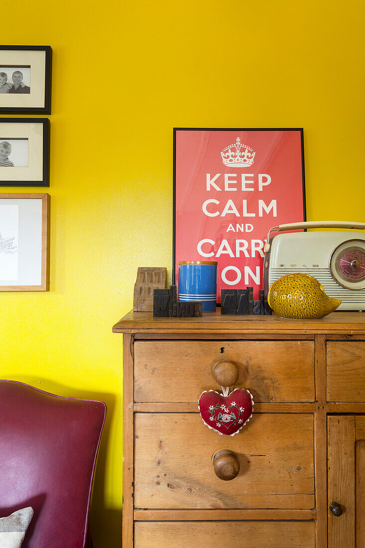 Yellow wall with Keep Calm and Carry On poster above antique wooden chest of drawers with decorative items
