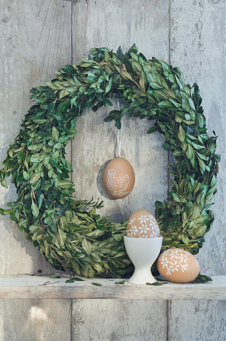 Painted Easter eggs, egg cup and box tree wreath on shelf in front of wooden wall