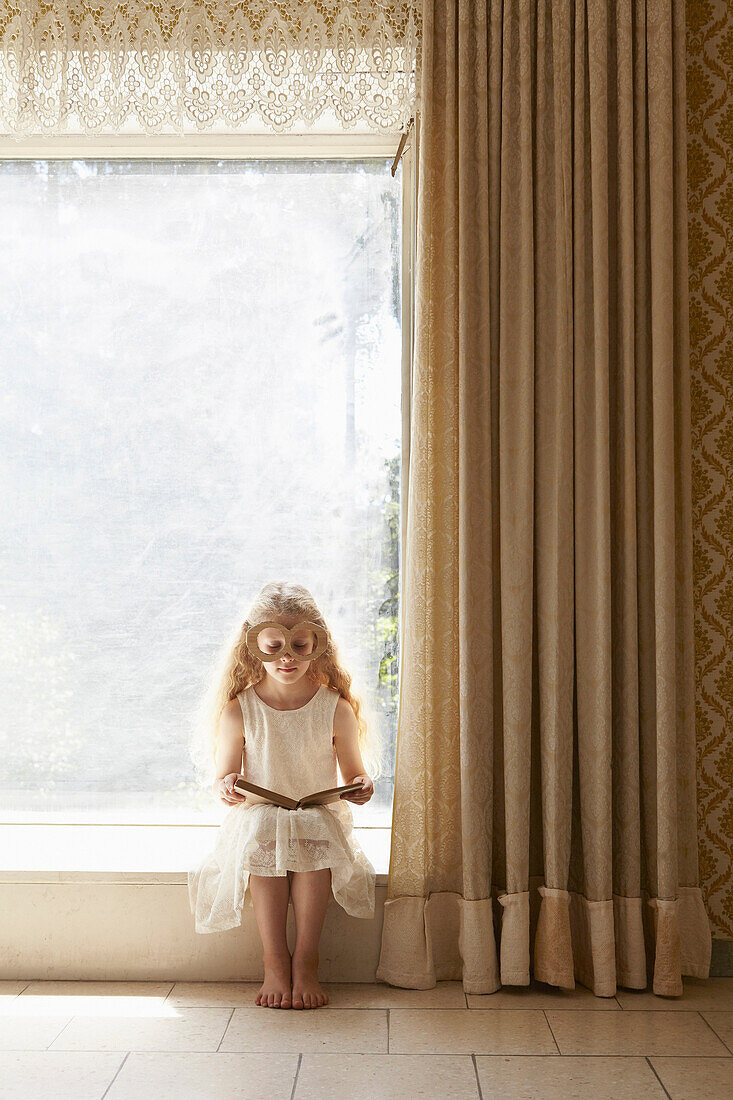 Girl sitting on windowsill wearing cardboard glasses reading book