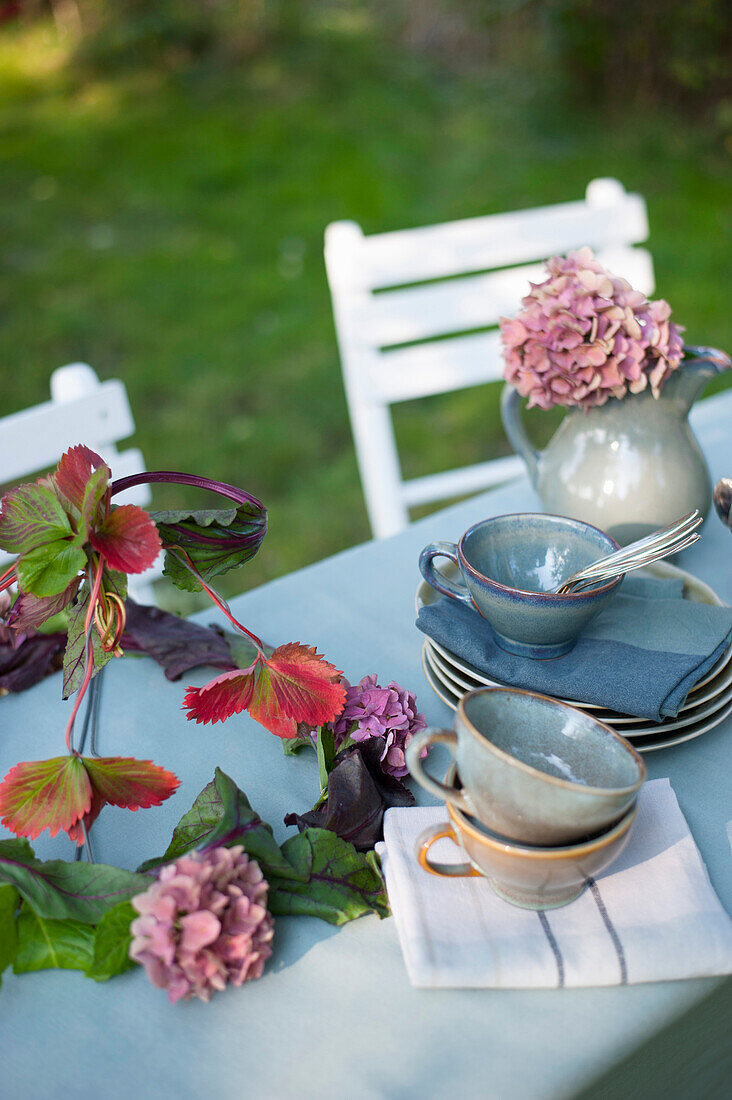 Geschirr und DIY-Lampenschirm aus Blättern und Hortensienblüten, die auf einem Couchtisch im Garten liegen