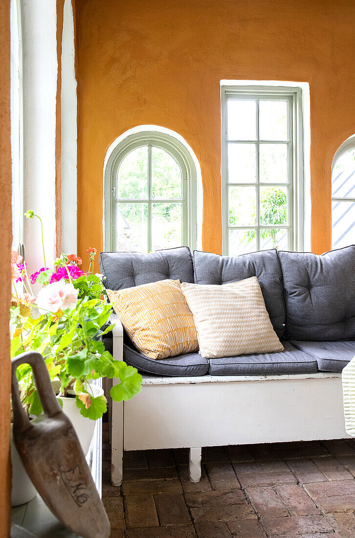 Holzbank mit Kissen in der Orangerie mit teilweise gewölbten Fenstern und ockergelben Wänden
