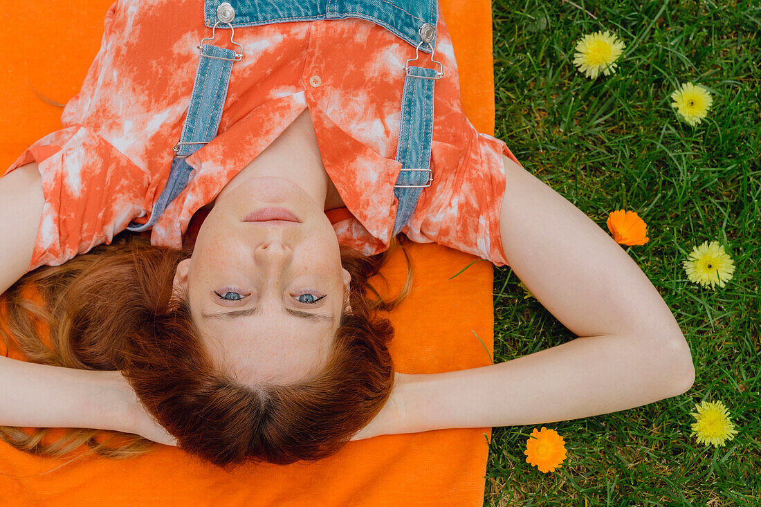 Relaxed young woman lying with hands behind head on picnic blanket by flowers at back yard