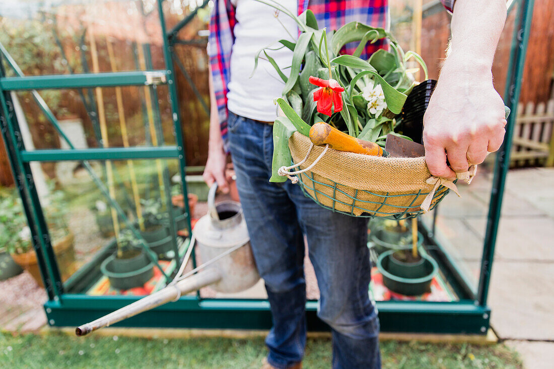 Nahaufnahme eines Mannes mit Korb mit Blumen und Gießkanne vor dem Gewächshaus im Garten