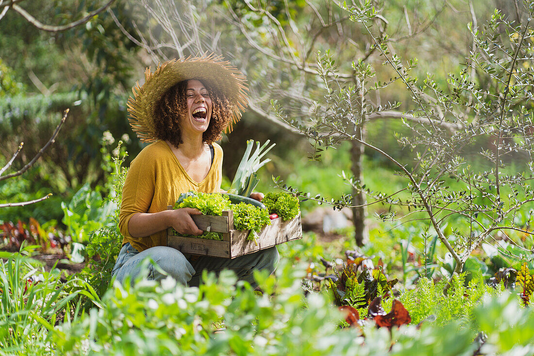 Laughing woman holding crate of green vegetables while squatting in garden